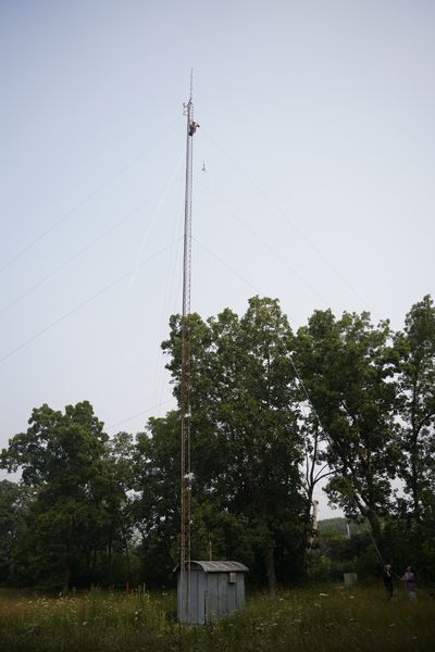 A guyed lattice tower in a field with a climber near the top and ground crew below